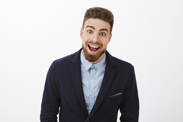 Disparo de cintura para arriba de un joven y carismático soltero rico entusiasmado, encantado y sorprendido, con un elegante traje con barba y ojos azules, sonriendo ampliamente de asombro y alegría mirando impresionado