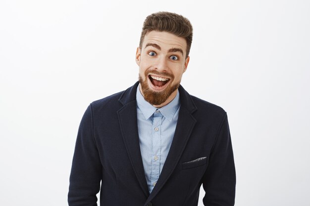 Disparo de cintura para arriba de un joven y carismático soltero rico entusiasmado, encantado y sorprendido, con un elegante traje con barba y ojos azules, sonriendo ampliamente de asombro y alegría mirando impresionado