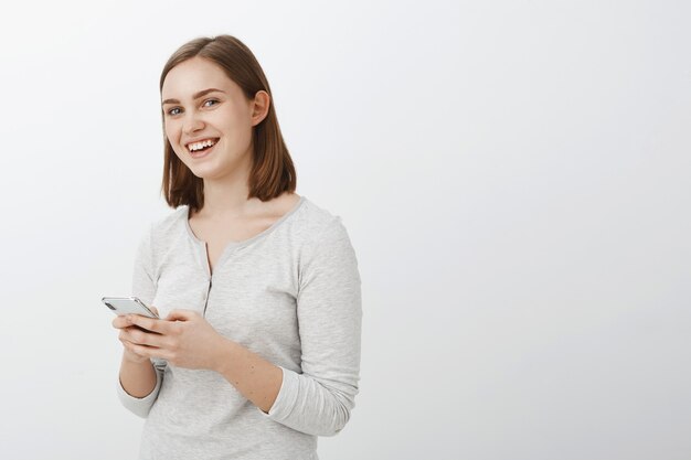 Disparo de cintura para arriba de una mujer morena de aspecto amistoso creativo y entusiasmado con una blusa casual de pie medio volteado sobre una pared gris sosteniendo un teléfono inteligente riendo entretenido y divertido