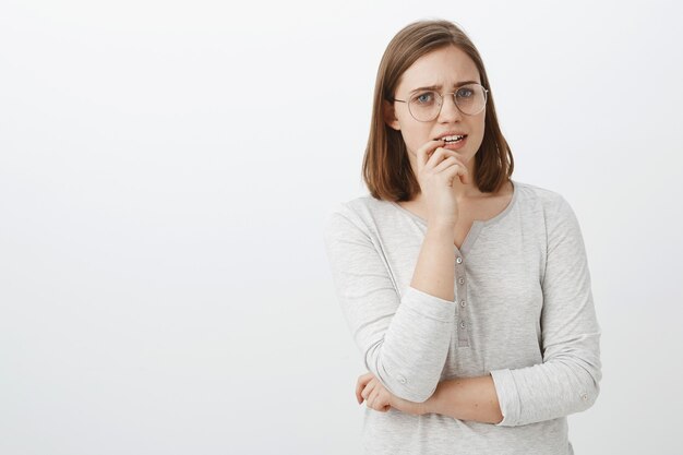 Disparo de cintura para arriba de preocupada mujer europea linda preocupada con gafas y blusa sosteniendo el dedo en el labio frunciendo el ceño con aspecto nervioso y preocupado esperando noticias del hospital sosteniendo la mano cruzada sobre el cuerpo