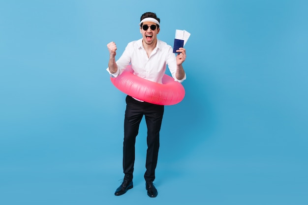 Disparo de cuerpo entero de hombre disfrutando de viaje de vacaciones. Chico en traje de negocios y gafas de sol con documentos, entradas y círculo inflable rosa.