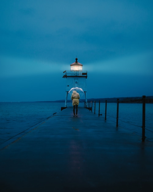 Foto gratuita disparo distante vertical de una persona sosteniendo un paraguas caminando sobre una pasarela cerca de un faro