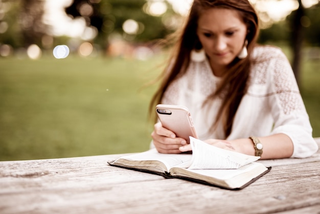 Disparo de enfoque selectivo de una mujer usando su teléfono inteligente con una Biblia abierta sobre la mesa