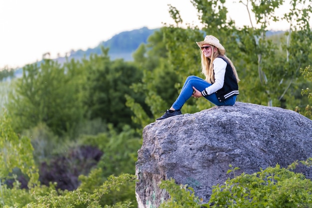 Disparo de enfoque superficial de una mujer europea con sombrero de vaquero sentado sobre una roca en la naturaleza