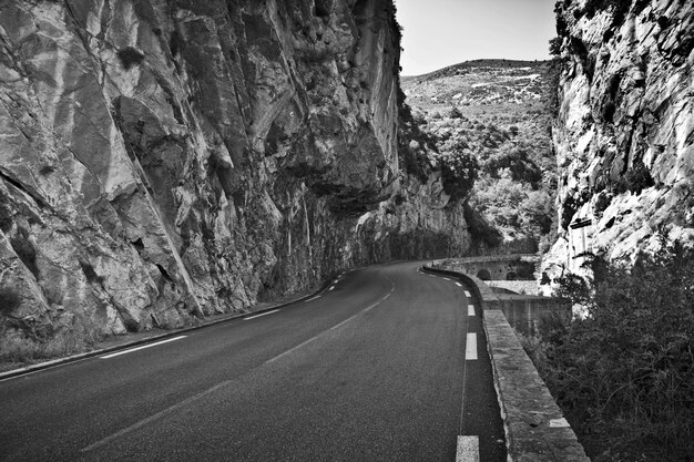 Disparo en escala de grises de una carretera vacía rodeada de rocas bajo la luz del sol durante el día