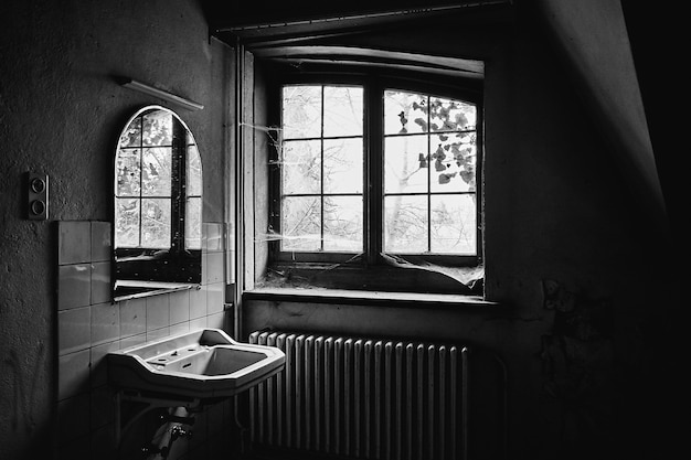 Disparo en escala de grises de una habitación abandonada con un fregadero y un espejo y telarañas por toda la ventana