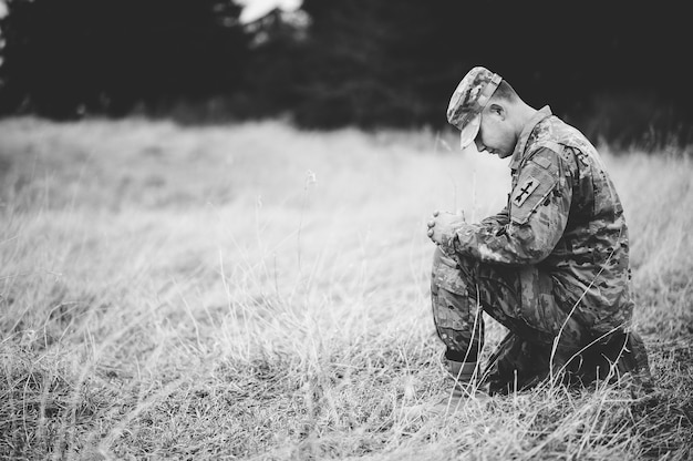 Disparo en escala de grises de un joven soldado rezando mientras está de rodillas sobre una hierba seca