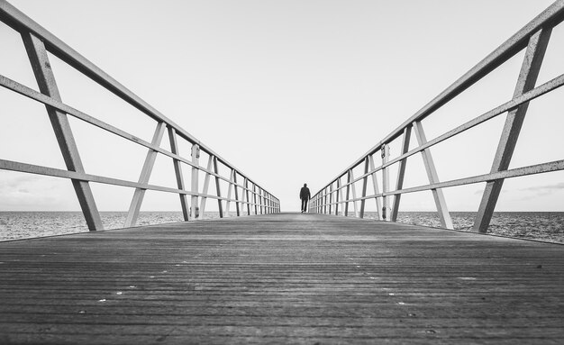 Disparo en escala de grises de una persona caminando sobre un puente de madera junto al océano - concepto: salida, separación