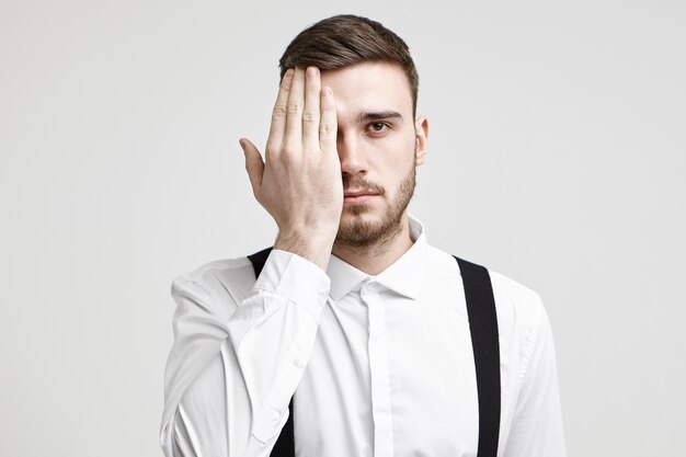 Disparo de estudio aislado de apuesto joven trabajador corporativo con cerdas y elegante corte de pelo mirando a la cámara, cubriendo un ojo con la palma como si le hicieran una prueba de ojos durante un examen de la vista