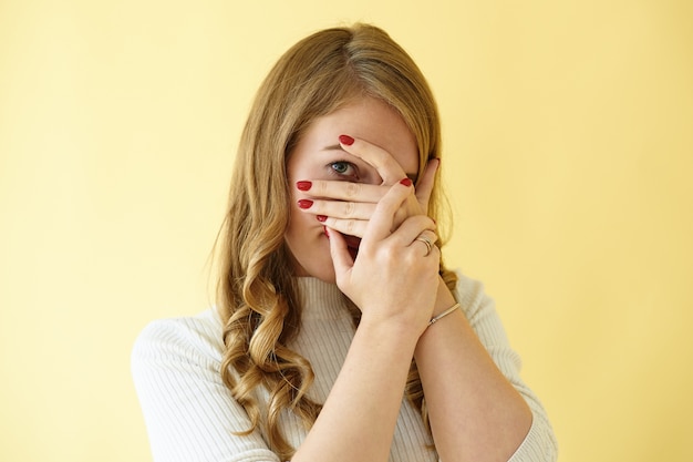 Disparo de estudio aislado de glamorosa mujer europea joven con estilo con uñas rojas cuidadas cubriendo su rostro con las manos y mirando a través de sus dedos a la cámara, sintiéndose tímida, avergonzada o asustada