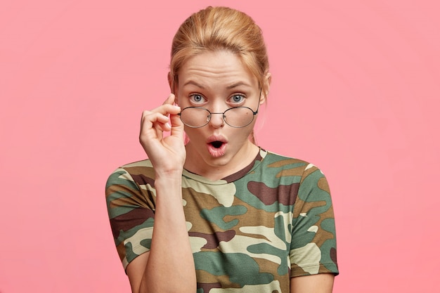 Disparo de estudio de atractivo modelo femenino joven mira a través de gafas redondas con expresión atenta y sorprendida, vestida con camiseta casual, aislada sobre rosa
