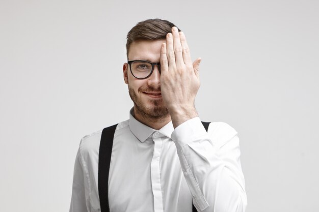 Disparo de estudio horizontal de feliz atractivo joven empresario barbudo con ropa formal y gafas que cubren la mitad de su rostro mientras le examinan los ojos en una cita de oftalmología