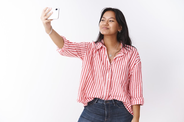 Disparo de estudio de una mujer asiática linda joven con estilo tomando selfie para impresionar a los seguidores en Internet con la nueva blusa de moda extendiendo la mano con el teléfono inteligente mirando la pantalla del teléfono móvil, fotografiando