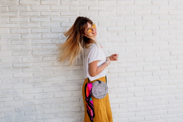 Disparo de estudio de mujer despreocupada en traje de verano brillante posando sobre pared de ladrillo blanco