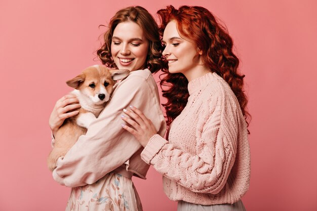 Disparo de estudio de niñas felices jugando con lindo perro. Riendo señoras caucásicas posando con mascota sobre fondo rosa.