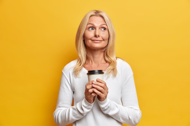 Disparo horizontal de la abuela reflexiva de ensueño tiene un descanso para tomar café disfruta del tiempo libre recuerda agradables recuerdos de su juventud posa con una taza de bebida caliente vestida de manera informal