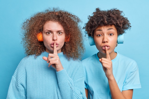 Disparo horizontal de una adolescente diversa que dice que se quede callada, que haga un signo de silencio, que mantenga la información en secreto, que se vea misteriosamente vestida, que escuche música informal a través de auriculares aislados sobre una pared azul