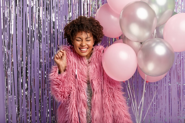 Disparo horizontal de la alegre dama afro aprieta el puño, disfruta de la celebración, vestida con un traje elegante, sostiene globos