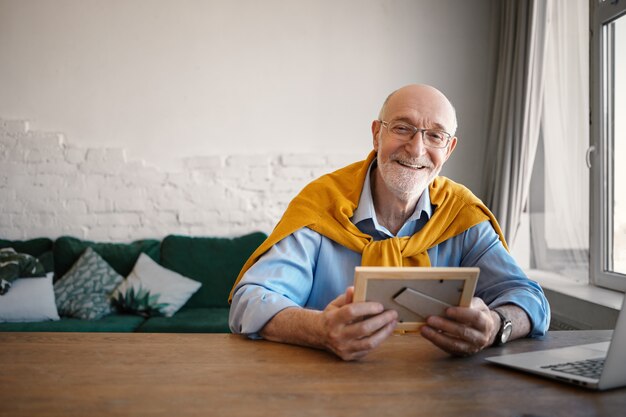 Disparo horizontal de alegre empresario de sesenta años con estilo con anteojos rectangulares sentado frente a una computadora portátil abierta, sosteniendo un retrato de familia en el marco de fotos y sonriendo felizmente