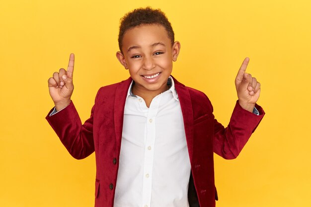 Disparo horizontal de alegre enérgico colegial afroamericano sonriendo, levantando los dedos, apuntando hacia arriba, indicando la pared del estudio en blanco con espacio de copia para su contenido publicitario