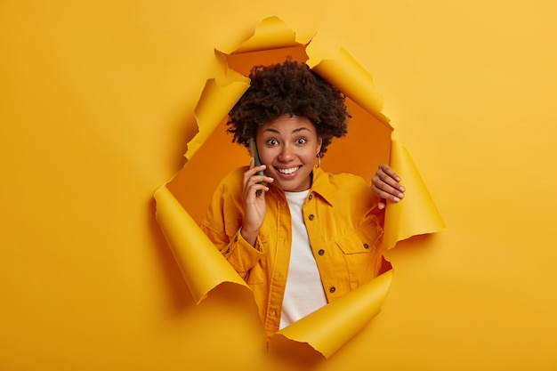 Disparo horizontal de alegre mujer afroamericana tiene conversación por teléfono celular, charla alegremente