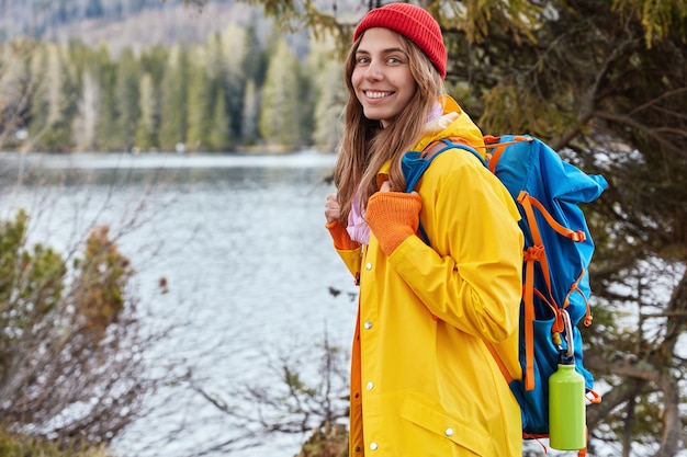 Disparo horizontal de alegre mujer caucásica vagabundo está de lado a la cámara, disfruta de la hermosa naturaleza