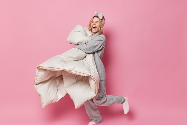 Disparo horizontal de una alegre mujer europea vestida con pijama que usa diadema se ríe alegremente sostiene un edredón suave que se divierte después de despertar aislado sobre un fondo rosado. Chica positiva con manta.
