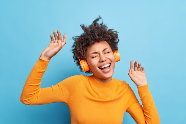 Disparo horizontal de alegre mujer de pelo rizado baila con el ritmo de la música disfruta de su lista de reproducción favorita, lleva auriculares estéreo en los oídos, capta cada bit canta una canción aislada sobre una pared azul