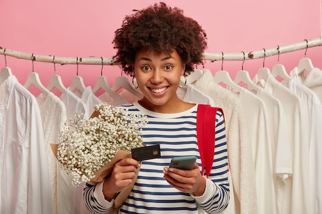 Disparo horizontal de alegre niña de pelo rizado sonríe agradablemente, utiliza un dispositivo moderno para pagar en línea, tiene tarjeta de crédito bancaria