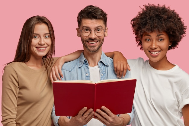 Disparo horizontal de alegres compañeros de grupo de raza mixta tres amistosos que están juntos de cerca, sostienen el libro de texto rojo, leen información para tareas comunes, aisladas sobre una pared rosa. concepto de educación.