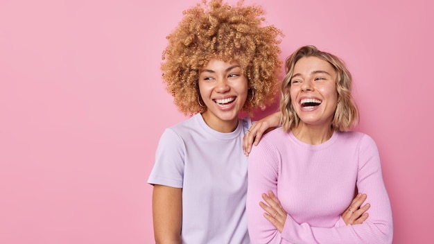 Foto gratuita disparo horizontal de alegres hermanas jóvenes que se ríen alegremente, se divierten, tienen una buena relación, miran hacia otro lado a algo divertido vestido casualmente aislado sobre un fondo rosa, copia en blanco, espacio para la promoción