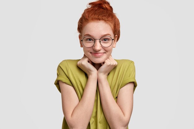 Disparo horizontal de atractiva mujer pelirroja que mantiene ambas manos debajo de la barbilla, se siente complacida al recibir felicitaciones, vestida con ropa informal, gafas redondas, aisladas sobre una pared blanca