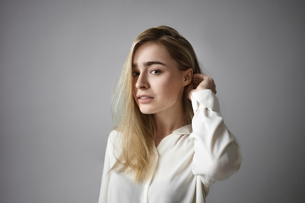 Disparo horizontal de atractiva mujer rubia en camisa blanca formal ajustando su peinado suelto y mirando a cámara con sonrisa pensativa. Personas, estilo de vida, expresiones faciales humanas y moda.