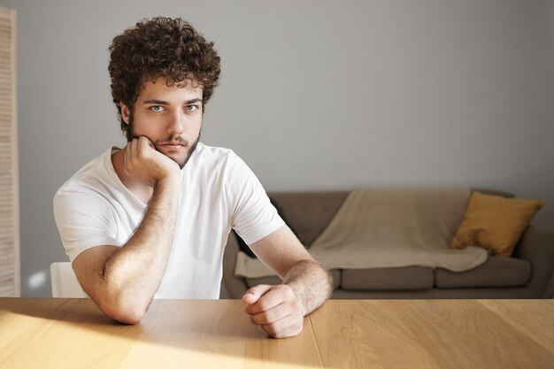 Disparo horizontal de atractivo joven serio con rastrojo habiendo aburrido la expresión facial, pasando el fin de semana solo en casa, sentado en un escritorio de madera contra la pared blanca,