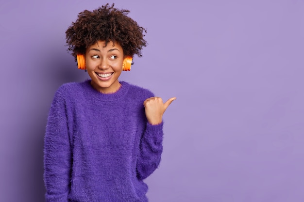 Foto gratuita disparo horizontal de buena apariencia alegre mujer afroamericana escucha pistas de audio apunta con el pulgar hacia afuera en el espacio vacío sonríe positivamente plantea