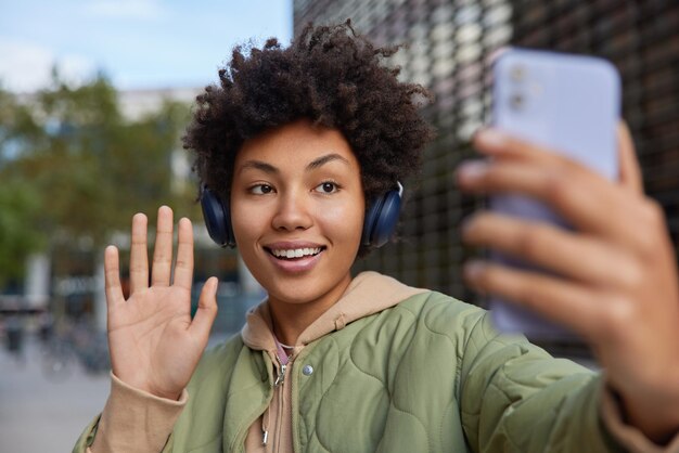 Disparo horizontal de buena mujer alegre con pelo rizado ondas palma en gesto de saludo saluda a mejor amigo utiliza auriculares estéreo