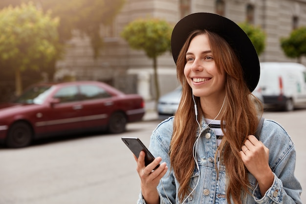 Disparo horizontal de chica alegre con sombrero de moda, pasa tiempo libre al aire libre