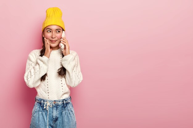 Disparo horizontal de una chica asiática guapa hace una llamada telefónica, disfruta de una conversación agradable a través del teléfono celular, vestida con ropa casual, se encuentra en el interior sobre un fondo rosa