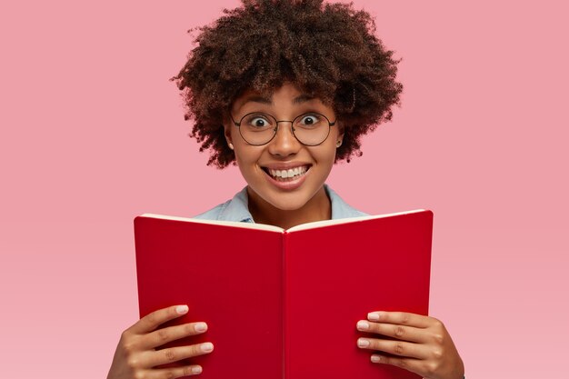 Disparo horizontal de una chica muy sonriente con una expresión facial agradable, lleva un libro de texto, usa gafas, se prepara para el examen, aislado sobre una pared rosa. Concepto de personas, etnia y alfabetización