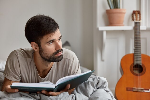 Disparo horizontal de un chico caucásico contemplativo con cerdas oscuras, viste ropa informal, se acuesta en la cama con un libro, se siente solo en casa, disfruta del fin de semana, tiene una vida tranquila. Ambiente doméstico, concepto de lectura