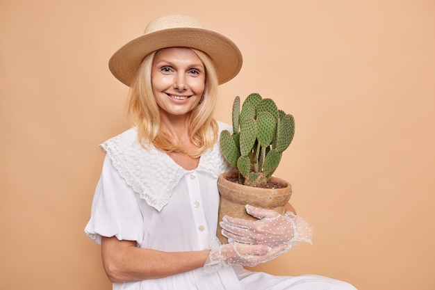 Disparo horizontal de una dama guapa con modales aristocráticos viste un atuendo elegante que lleva cactus en macetas a su jardín de casa sonríe complacida se sienta en el interior contra la pared marrón