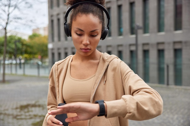 Disparo horizontal de una deportista seriamente motivada enfocada en el reloj inteligente revisa el tiempo sostiene una botella de agua fresca usa una sudadera con capucha marrón escucha música a través de poses de auriculares en la ciudad al aire libre durante el día