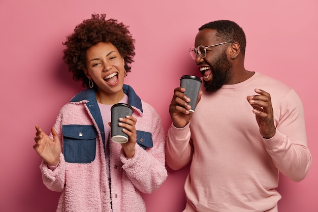 Disparo horizontal de divertida mujer alegre y hombre con piel oscura se divierten durante la pausa para el café, cantan canciones, bailan sin preocupaciones