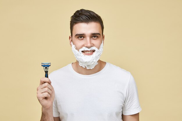 Disparo horizontal de divertido apuesto joven con espuma blanca en la cara con una sonrisa, sosteniendo el bastón de afeitar, yendo a afeitarse la barba, haciendo la rutina matutina. Aseo y belleza masculina