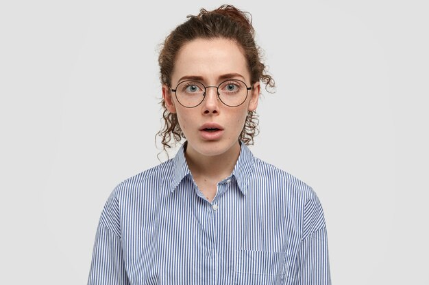 Disparo horizontal de estupefacta mujer pecosa con pelo rizado, lleva gafas redondas transparentes, elegante camisa a rayas