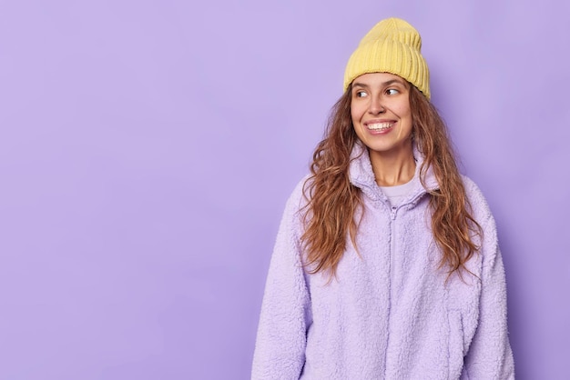 Disparo horizontal de feliz bonita adolescente sonríe positivamente mira hacia otro lado disfruta de un buen día viste un sombrero amarillo y una chaqueta de piel aislada sobre fondo púrpura espacio de copia para su contenido promocional