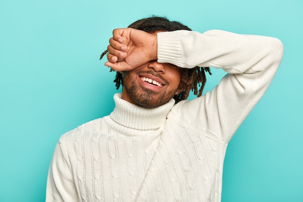 Disparo horizontal de feliz hombre afro con rastas, cubre los ojos, esconde la cara con el brazo, sonríe alegremente, viste un jersey blanco, posa sobre un fondo azul.