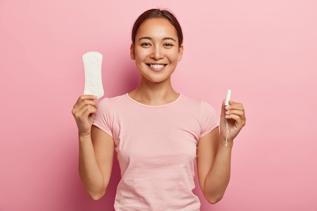 Disparo horizontal de feliz mujer coreana sostiene una toalla sanitaria y un tampón, demuestra productos íntimos para la salud de la mujer, sonríe suavemente, vestida con ropa informal, tiene días críticos.