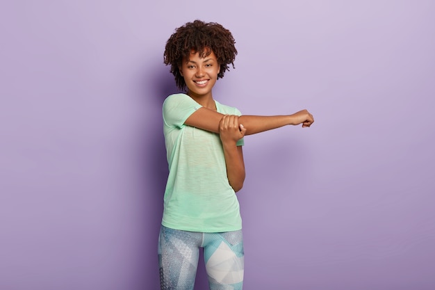 Disparo horizontal de feliz mujer deportiva afroamericana estira las manos antes del entrenamiento, sonríe alegremente, vestida con ropa deportiva, tiene cuerpo flexible