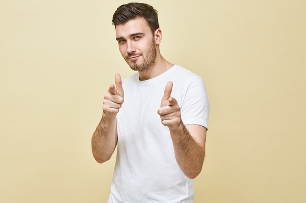 Disparo horizontal de guapo carismático joven con cabello negro y barba incipiente posando aislado apuntando con los dedos delanteros, con expresión facial coqueta segura, eligiéndote. Lenguaje corporal